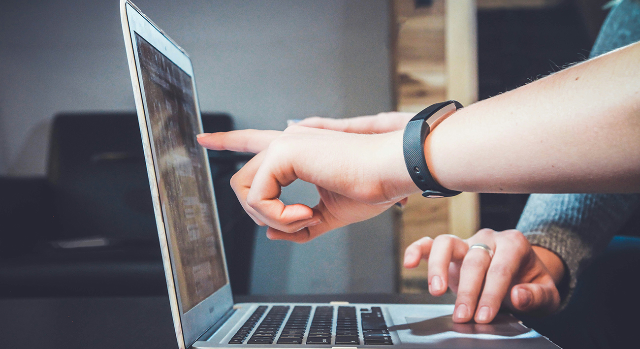Finger pointed to the screen on a laptop with another set of hands typing, signifying that one person is helping another and they have found what they needed.